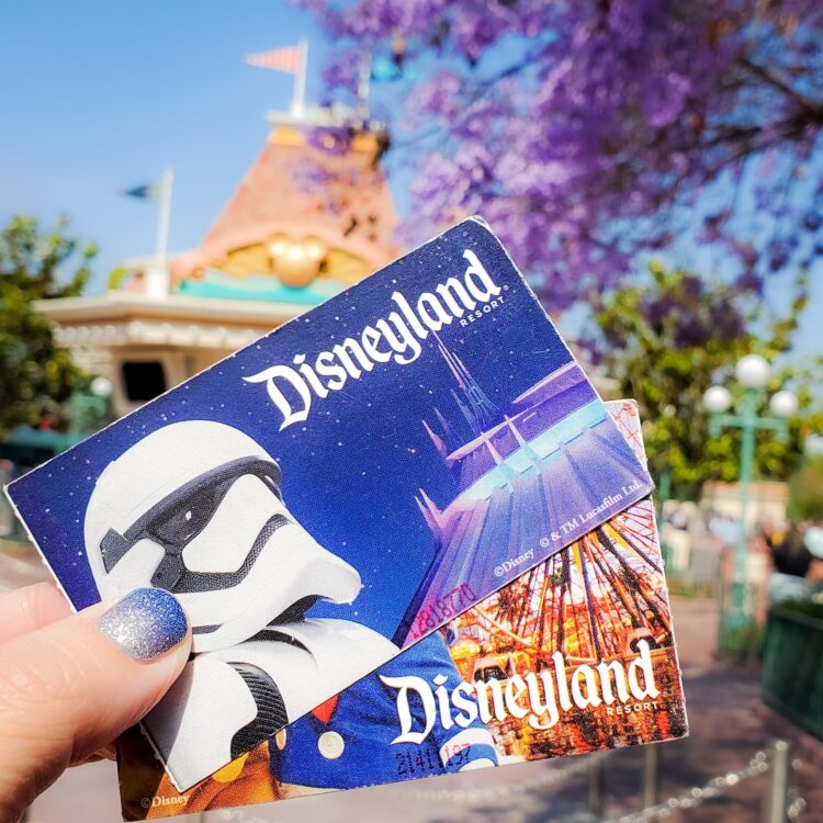 holding two Disneyland tickets , spread out, with Star Wars storm trooper on it, Disneyland ticket booth in background, purple blooms on tree