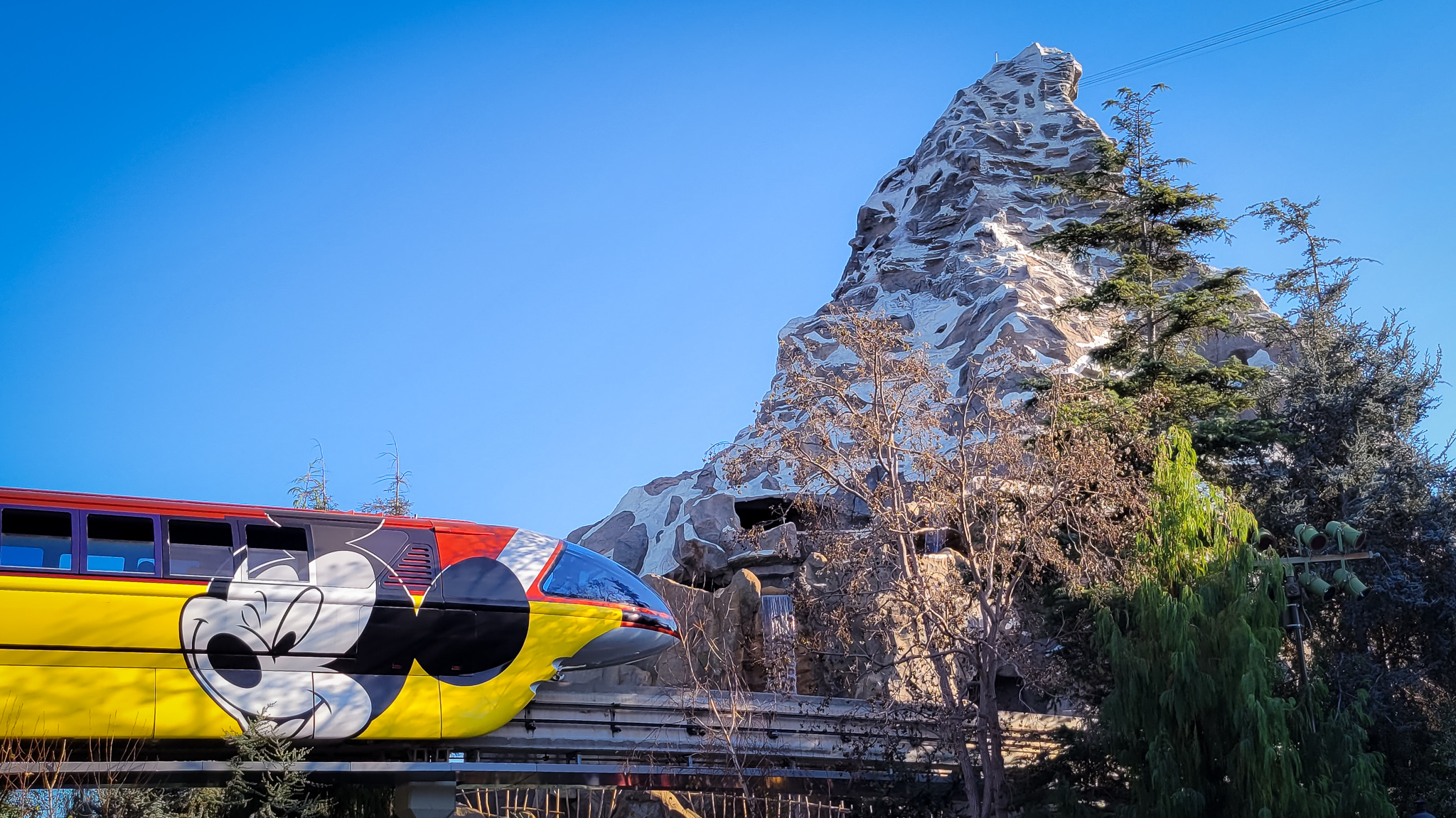 Monorail going past the Matterhorn at Disneyland. Mickey Mouse is on a red and yellow wrapped Matterhorn. Blue skies and a mixture of evergreen trees and trees bare with no leafs are seen.