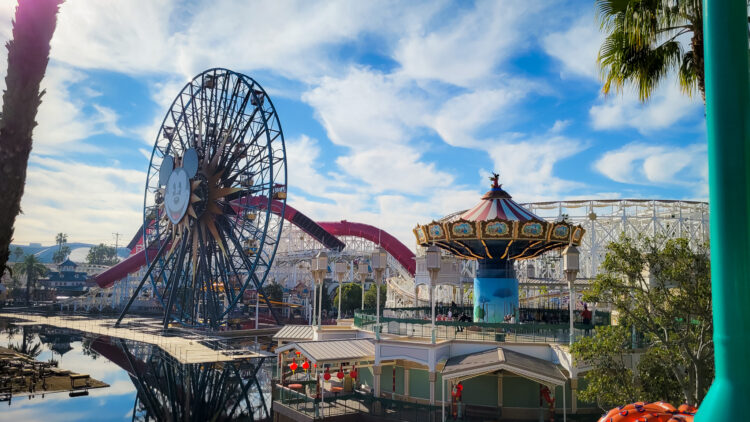 A view of Pixar Pier from across the bay, and while aflight a rides, taken from about 3 stories up. You see a ferris wheel with Mickey on it, Swings, and the Incredicoaster. Blue skies have white clouds passing 
