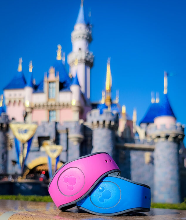 A pink and blue MagicBand sit on a ledge, with Sleeping Beauty Castle in the background. MagicBands are coming to Disneyland in 2022.