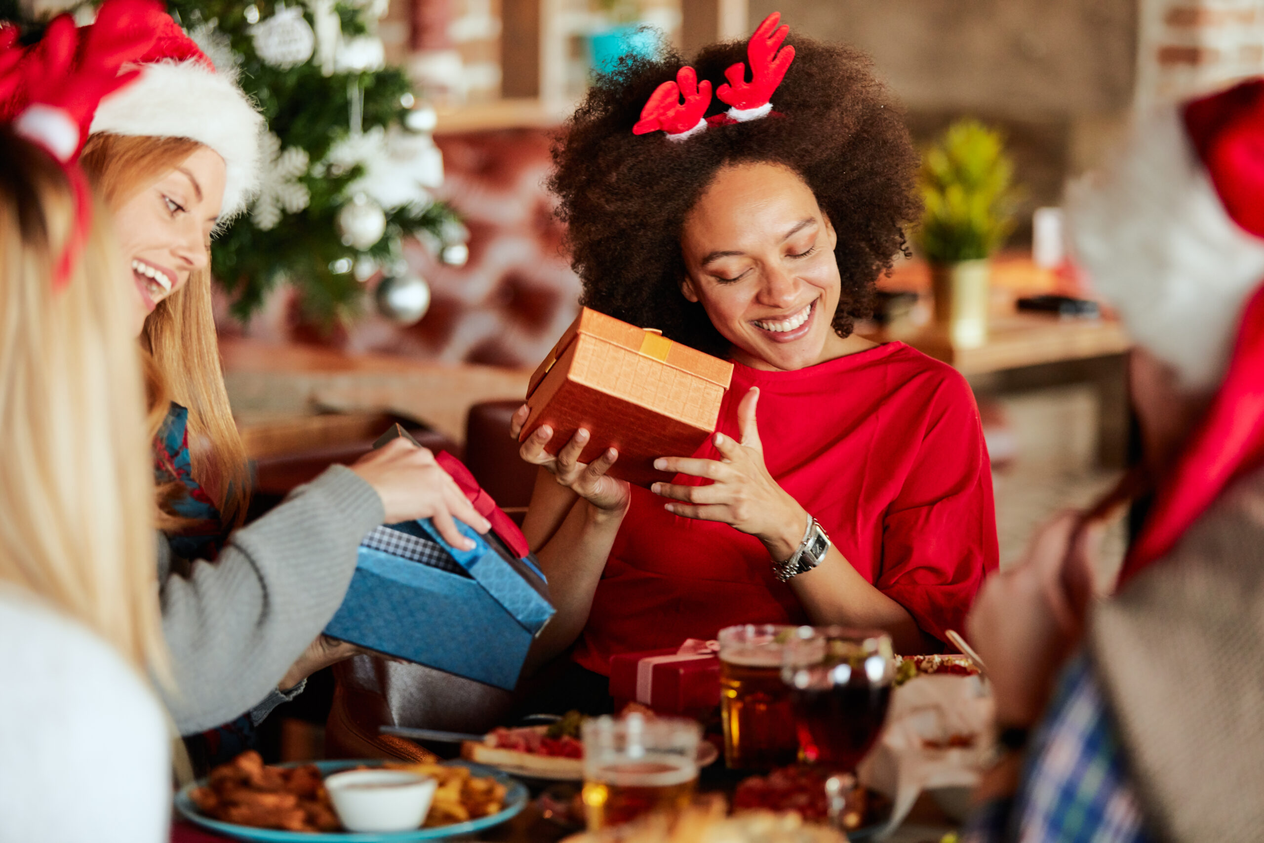 Women at a table exchanging and opening gifts while dresses in Christmas attire.