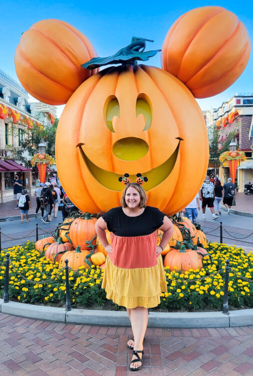Becca, dressed in a blank, orange and yellow dress with pumpkin Mickey ears in front of the Mickey Pumpkin on Main Street U.S.A. at Disneyland.