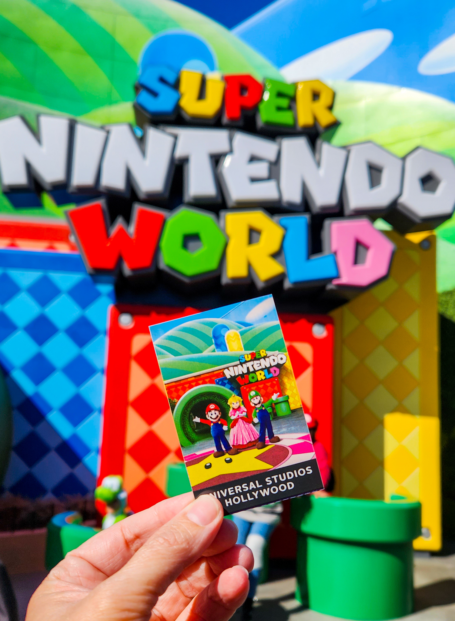 Hand holding a Universal Studios Ticket with Super Mario on it, in front of the actual entrance to Super Nintendo World in Universal Studios Hollywood