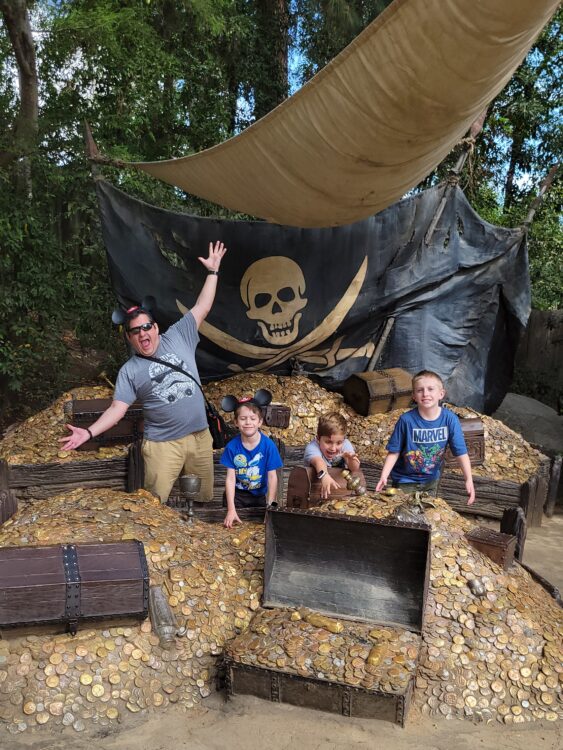 Becca's family enjoying the treasure they found on Pirates Lair on Tom Sawyer Island. Treasure chests and heaps of gold coins surround them with a Jolly Roger flag as a backdrop.